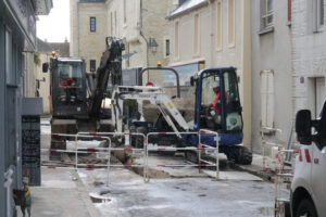 Travaux publics dans une rue étroite avec deux excavatrices en action, ouvriers en tenue rouge, bâtiments anciens en pierre en arrière-plan